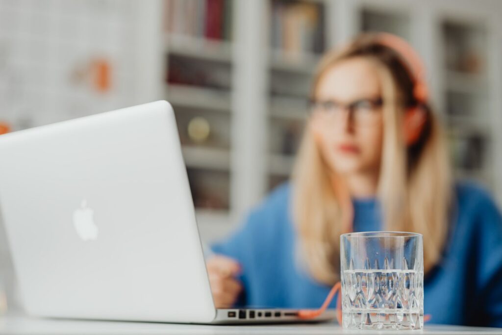 Laptop und Wasserglas am Schreibtisch, im Hintergrund eine konzentriert arbeitende Frau